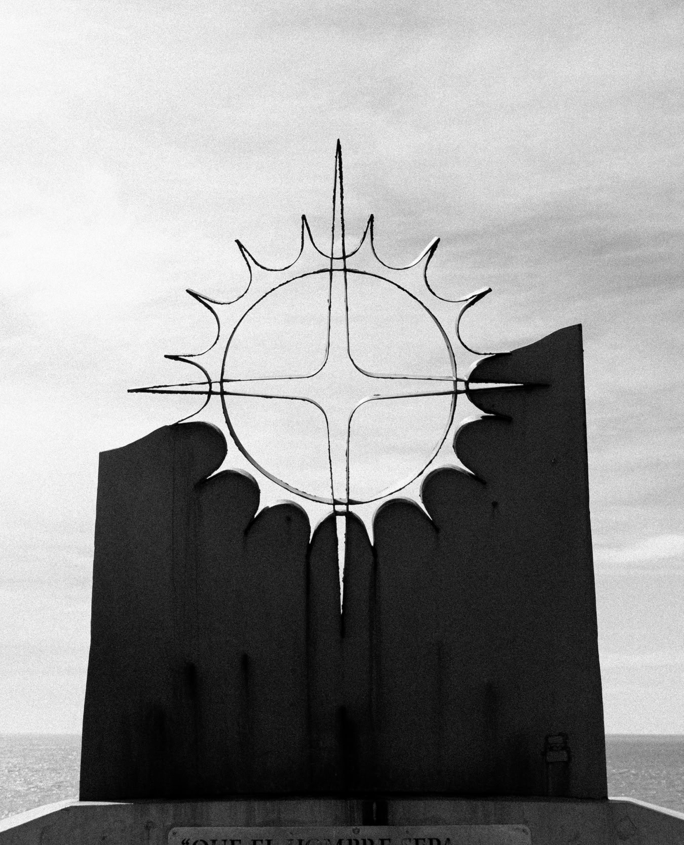 Schwarzweißfoto einer abstrakten Skulptur mit Sonnenmotiv vor einer Küstenkulisse.