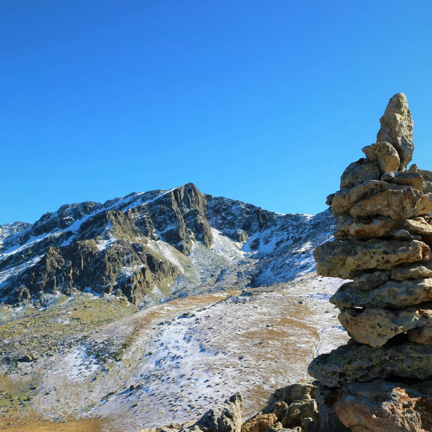 Ruhige Berglandschaft mit einem Steinhaufen unter einem klaren blauen Himmel und schneebedeckten Gipfeln.